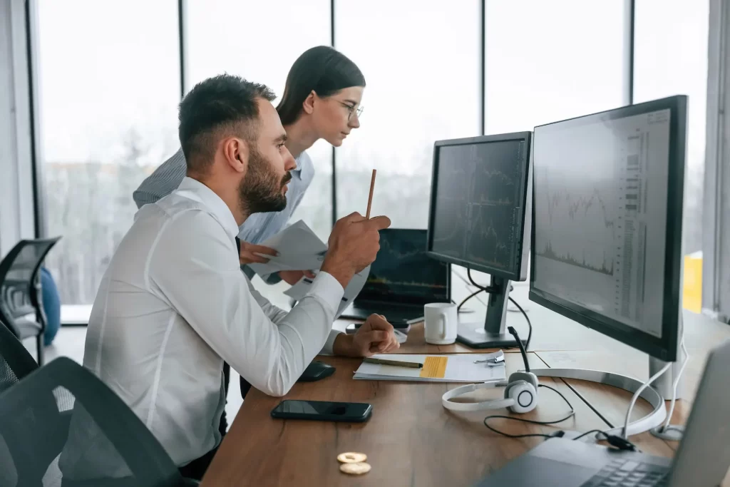 A man and a woman are working together assessing the business finances and cash flow of the company at his desk with charts and reports on the screens