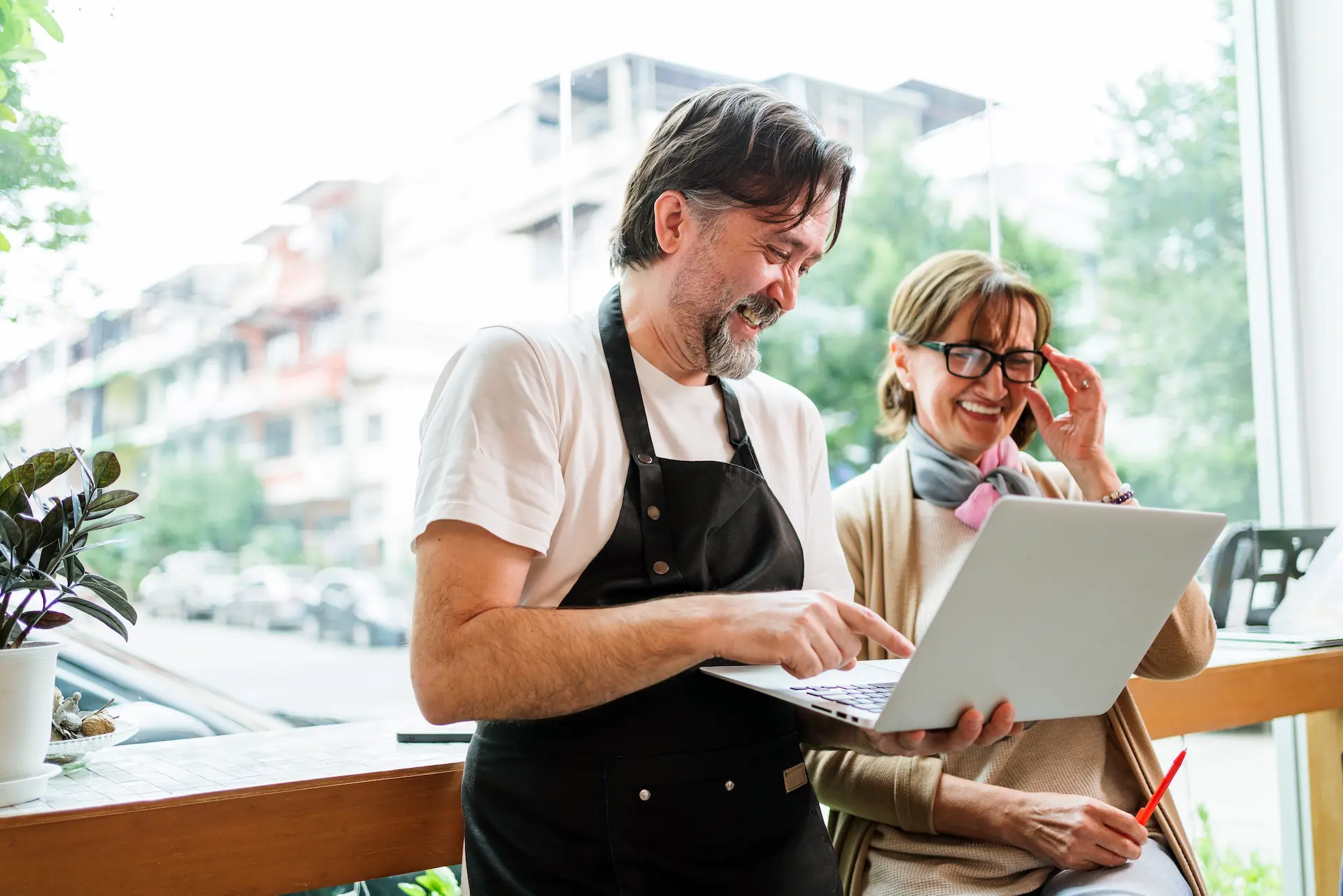 A smiling male business owner points at a laptop, showing a laughing female customer the screen. They are in a cafe.