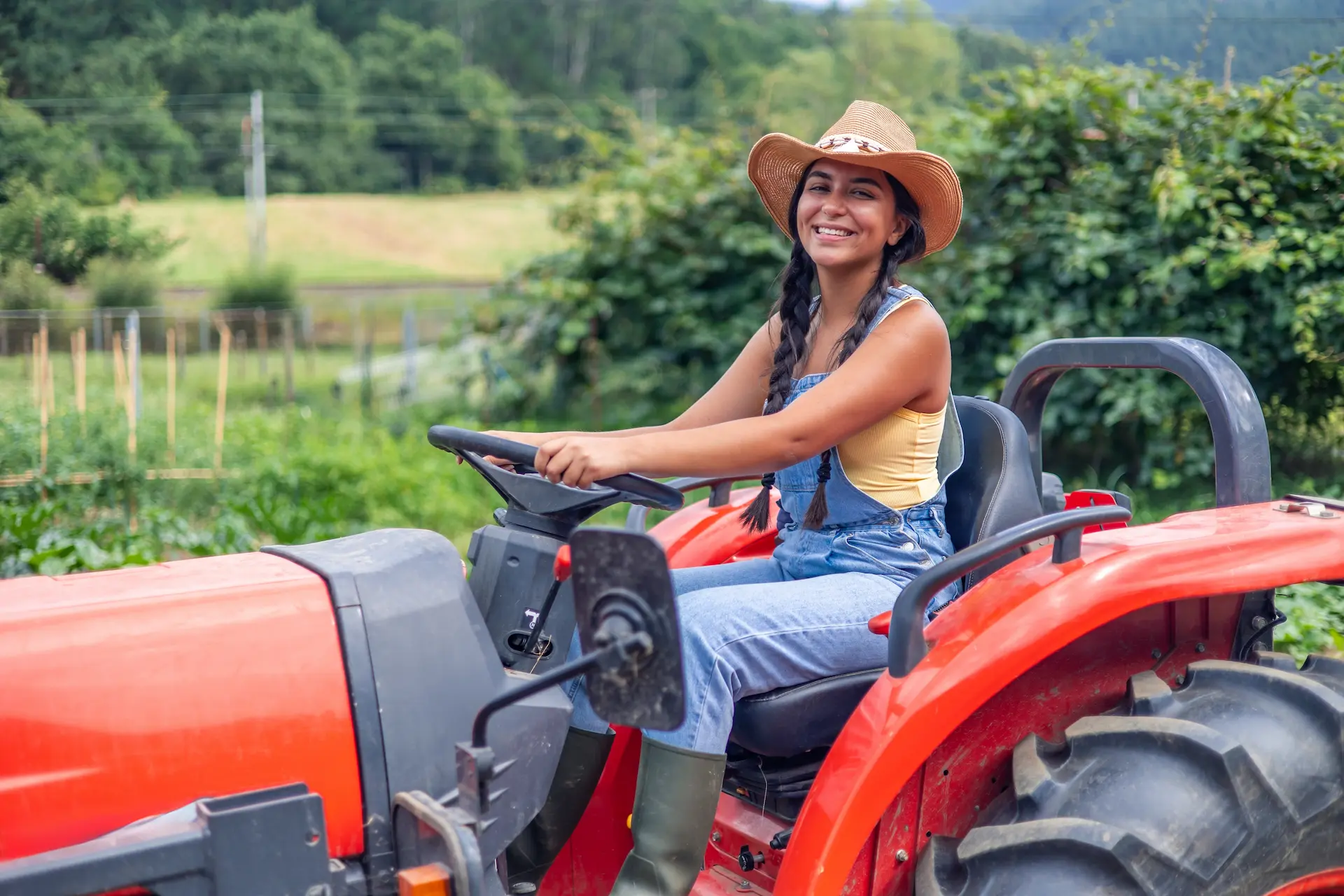 Young woman farmer wearing a hat and overalls, smiling while driving a bright red tractor through a lush cultivated field on a sunny summer day in the countryside